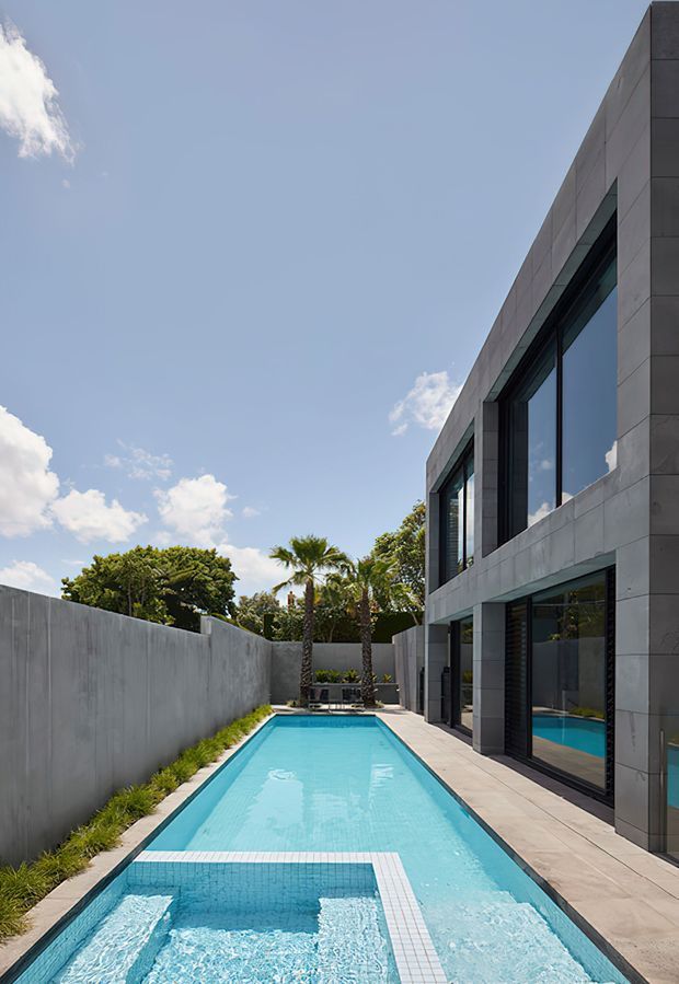 Exterior architectural photograph of a sleek modern two-storey residence beside a long narrow swimming pool. The house has a rectilinear form, dark-framed floor-to-ceiling glazing, and large grey concrete or stone-look facade panels. A raised spa or water feature occupies the foreground, with a paved poolside walkway, tall privacy wall, and tropical planting visible around the courtyard.