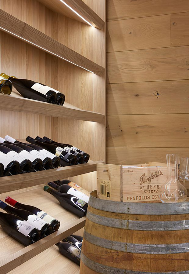 Close-up interior view of a compact wine storage area, with extensive light timber shelving, integrated linear LED lighting, horizontal timber wall boards, wine bottles, and a rustic wooden wine barrel used as a display surface. The space has a warm, refined cellar-like atmosphere.