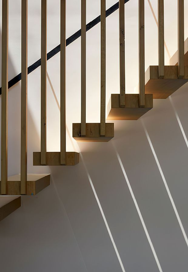 Close-up architectural photograph of a modern open-riser staircase. Thick timber treads project from a pale wall, with slim vertical timber balustrade members extending upward. Strong sunlight creates diagonal linear shadows across the wall, emphasizing the sculptural stair geometry and minimalist detailing.