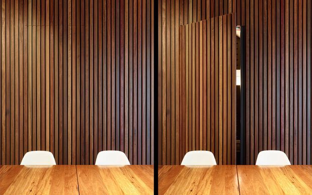 A close-up interior view of a dining area with a warm timber table in the foreground, white dining chairs, and a dramatic full-height vertical timber slat wall behind. A narrow opening in the slatted wall reveals a darker adjoining space. The composition is symmetrical and strongly emphasizes natural wood texture and vertical rhythm.