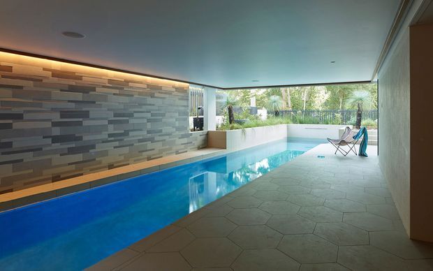 A refined indoor swimming pool area with a long narrow pool, geometric stone-tile flooring, a layered horizontal stone feature wall with concealed linear lighting, and broad glazing opening toward landscaped greenery. Minimal lounge seating sits at the far end, creating a calm, resort-like atmosphere.