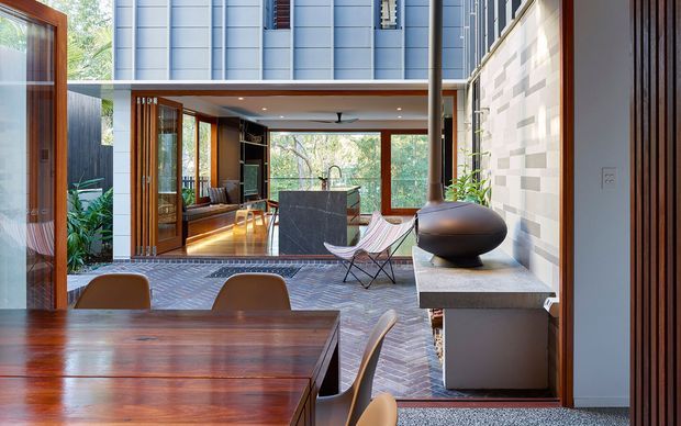 Contemporary open-plan interior combining a dining area, kitchen and living space. The room features a substantial dark timber dining table, warm timber-framed glazing and doors, a dark stone kitchen island, a sculptural freestanding fireplace on a stone ledge, and a patterned light brick or tile feature wall. Large windows provide views to surrounding greenery.