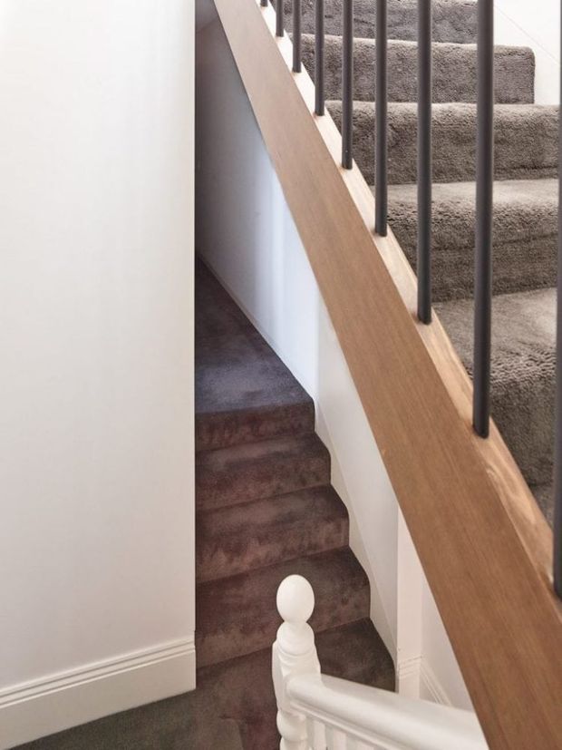 Interior view of a carpeted staircase turning upward, with a dark brown carpet runner, white painted walls and skirting, a timber handrail, and slim dark metal balusters. A traditional white stair newel is visible in the foreground.
