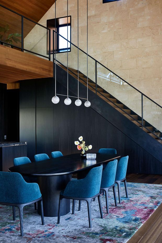 A sophisticated dining area beneath a dramatic staircase and mezzanine. The room features a dark oval dining table with upholstered teal-blue chairs, globe pendant lights, black built-in cabinetry, warm timber ceilings and flooring, a pale stone masonry wall, and a colourful patterned rug. A black-framed glass balustrade and stair structure create a strong contemporary architectural focal point.