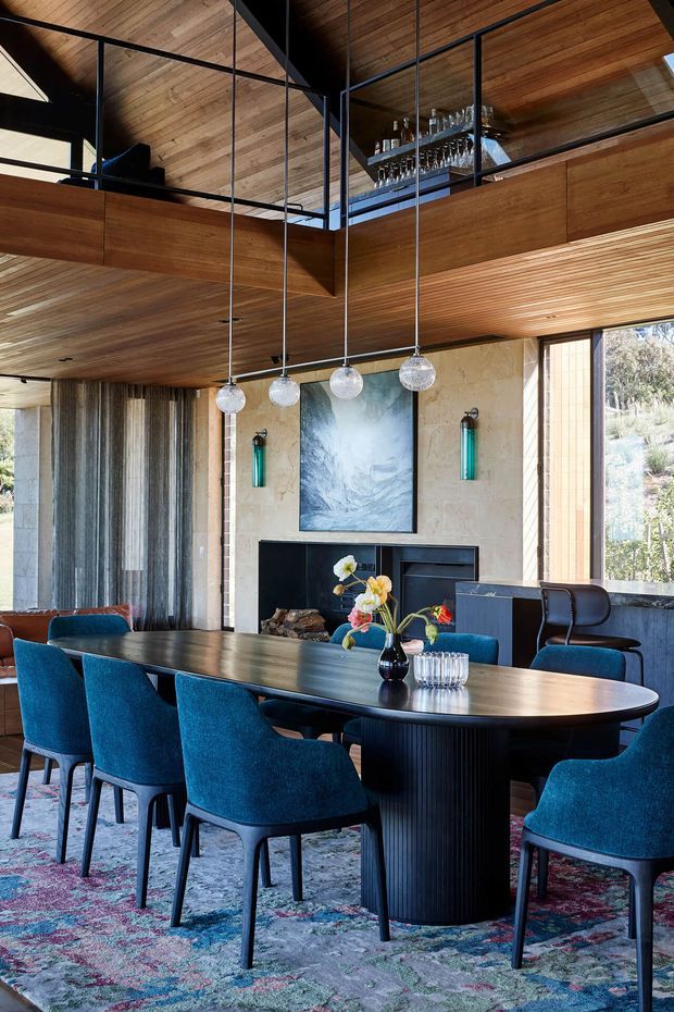 A sophisticated dining room with a large oval dark timber dining table surrounded by deep blue upholstered chairs. The room features extensive warm timber wall and ceiling lining, black-framed glass balustrades overlooking an upper level, sculptural pendant lights, a stone fireplace wall with artwork, and large windows connecting to the natural landscape outside. The interior has a refined, dramatic character with strong mid-century-inspired detailing.