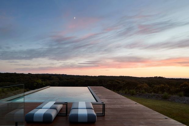 A tranquil architectural rooftop or elevated deck overlooking a wooded landscape at sunset. A long rectangular infinity-edge pool is integrated into a broad timber deck, with two striped cushioned sun loungers and clear glass balustrading in the foreground. The sky is softly coloured with pink, orange, and blue tones, creating a serene resort-like outdoor setting.