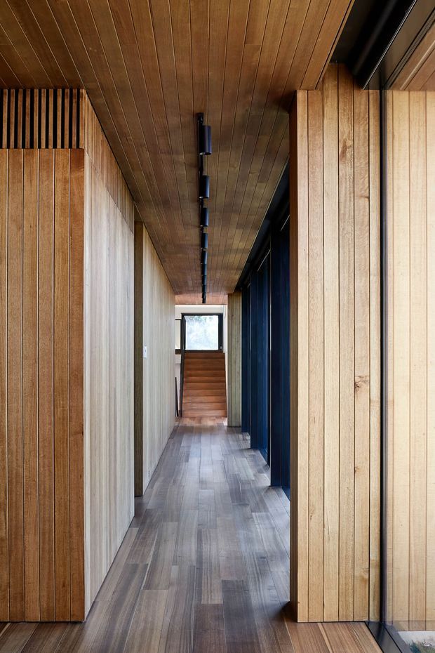 A long, narrow interior corridor lined with extensive vertical timber wall panelling and a matching timber-plank ceiling. Grey-brown plank flooring runs toward a short staircase and bright window at the far end. Dark blue-black door or wall panels contrast with the warm wood, while a linear row of black recessed or surface-mounted lights follows the ceiling. The space has a refined, contemporary timber-lined appearance.