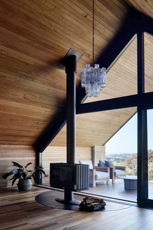A dramatic, timber-lined living room with a high angular ceiling and expansive black-framed glazing. A suspended crystal chandelier hangs beside a freestanding black wood-burning stove and television, while a lounge area opens onto a timber deck with distant landscape views. The space combines warm natural timber, dark structural framing, and contemporary furnishings.