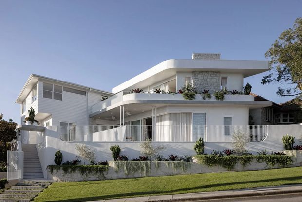 Exterior view of a bright white multi-level residence on a sloping suburban site. The house features a broad curved flat roof, rounded balcony and terrace edges, white horizontal cladding, pale masonry feature walls, extensive white-framed glazing, vertical privacy screens, planted balcony beds, and stepped landscaped retaining walls.