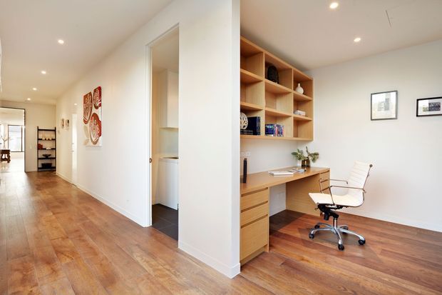 Bright open-plan home office nook integrated into a white-walled hallway, with a built-in timber desk, drawers, and open shelving. The space has warm timber flooring, recessed ceiling lights, framed artwork, and a white swivel office chair. A utility or storage room is visible through the adjacent doorway.