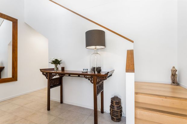 Bright residential entry hall featuring a dark carved timber console table, large wood-framed mirror, tiled flooring, white walls, a contemporary pendant lamp, and timber steps leading to a raised area. Traditional furniture contrasts with a clean modern interior shell.