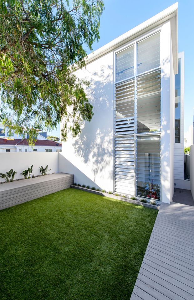 Bright contemporary courtyard beside a white modern house, featuring a compact green lawn, grey timber decking, a built-in timber bench, and a tall glazed facade with prominent horizontal louvres. A leafy tree provides partial shade, while white walls and clean geometric lines create a crisp outdoor setting.
