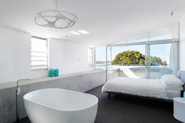 Bright contemporary bedroom with an adjoining open bathroom area, featuring a freestanding white bathtub, concrete vanity or partition, dark carpeted floor, white walls, a sculptural pendant light, and extensive glazing overlooking water and greenery.