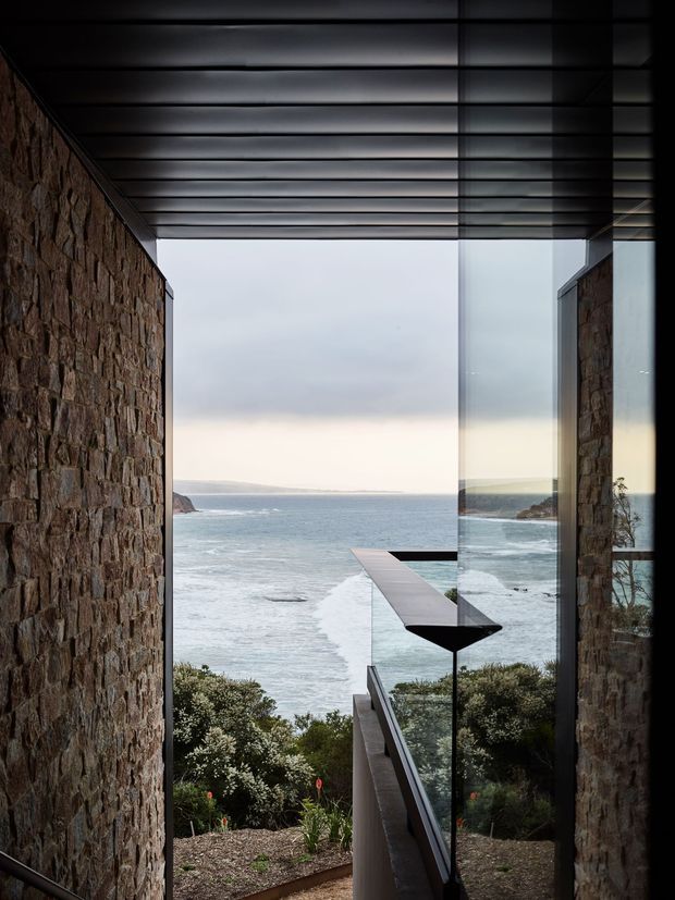 A dramatic coastal architectural passage framed by rough natural stone walls, dark metal roof beams, and floor-to-ceiling glazing. The view opens directly toward the ocean and rugged coastline, with a narrow projecting terrace or walkway extending toward the water. The composition emphasizes strong horizontal and vertical lines, reflective glass, and a moody, minimalist material palette.
