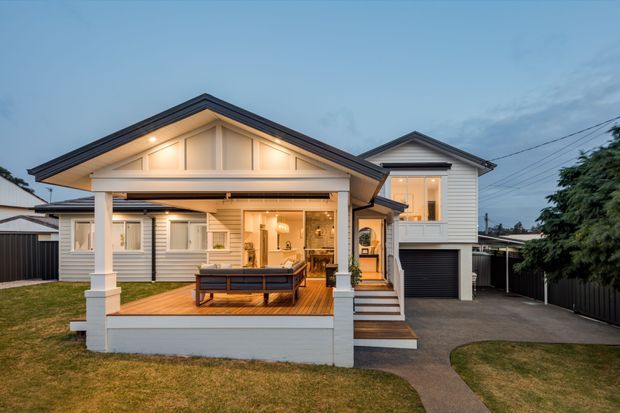 Exterior view of a renovated or newly built suburban home at dusk, featuring a light weatherboard facade, prominent gabled rooflines, a covered front veranda with white posts and detailed gable trim, warm exterior lighting, timber decking, and an attached single-car garage. The open veranda reveals a furnished outdoor seating area and glimpses of the warmly lit interior.