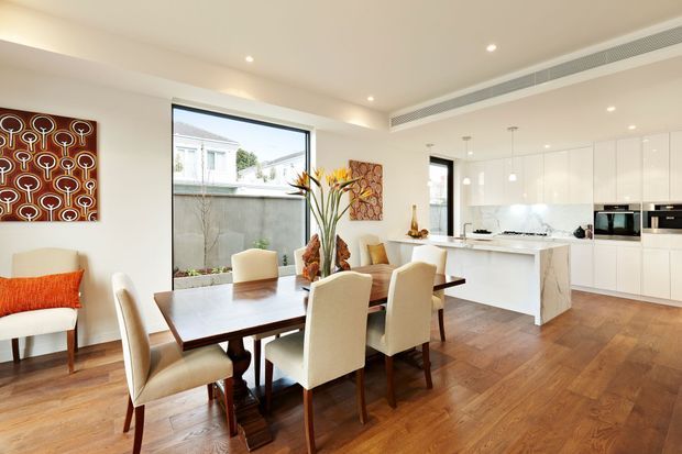 Bright open-plan dining and kitchen interior with a polished contemporary fit-out. A large timber dining table with upholstered chairs occupies the foreground, while a white island, glossy white cabinetry, integrated appliances, and pendant lights define the kitchen behind it. Warm timber flooring contrasts with white walls and joinery, and colourful artwork and tropical floral arrangements add decorative accents. Large windows provide natural light and views to an exterior courtyard.