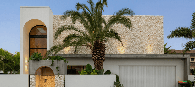 Contemporary two-storey residential facade with a flat roof, pale rendered walls, prominent rounded arch openings, a large vertically glazed entry feature, textured light natural-stone cladding, and an integrated garage. Tropical palm landscaping is positioned in front of the home.