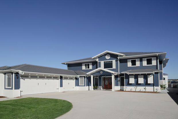 A large two-storey coastal-style residence with blue-grey horizontal cladding, white trim, a dark grey low-pitched roof, prominent gabled entry, circular attic window, and an attached multi-car garage. The home fronts a broad concrete driveway with a small lawn under a clear blue sky.