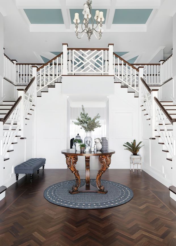 A bright, formal entry foyer with a dramatic symmetrical double staircase rising to a central landing. White painted balustrades and walls contrast with dark timber handrails, stair treads, and a richly patterned herringbone timber floor. A carved dark-wood console table, decorative objects, a round rug, bench, and chandelier complete the refined interior. The ceiling features recessed geometric panels with pale blue-green inserts.