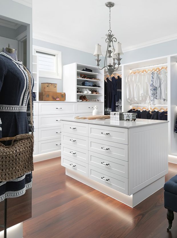 Bright, refined walk-in wardrobe with extensive white built-in drawers, open clothing rails, shelving for shoes and accessories, and a central island. The room features pale blue-grey walls, white trim, polished dark timber flooring, a decorative chandelier, and a dressed mannequin displaying clothing.