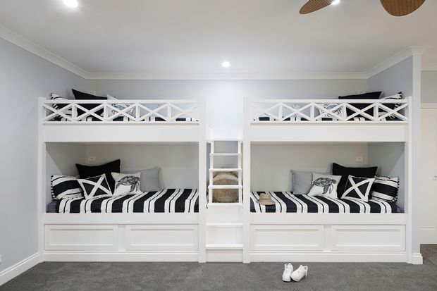 Bright, symmetrical children's bedroom featuring two built-in white bunk-bed units with decorative crisscross railings, integrated lower storage drawers, central shelving, and coordinated black-and-white striped bedding. The room has pale grey walls, white trim and cabinetry, recessed ceiling lights, and grey carpet.