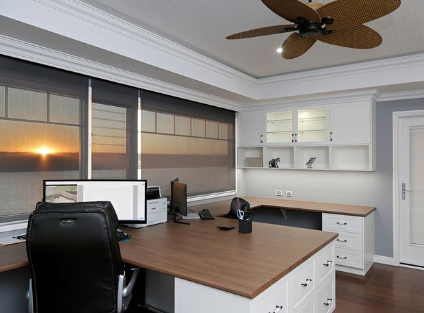 A bright, well-appointed home office with a large L-shaped timber worktop and white built-in cabinetry. Multiple computer workstations sit in front of broad windows with translucent roller blinds, while open shelving and drawers provide storage. The room features grey walls, white decorative cornices and wall panelling, a white tongue-and-groove ceiling, dark timber flooring, and a sculptural ceiling fan.