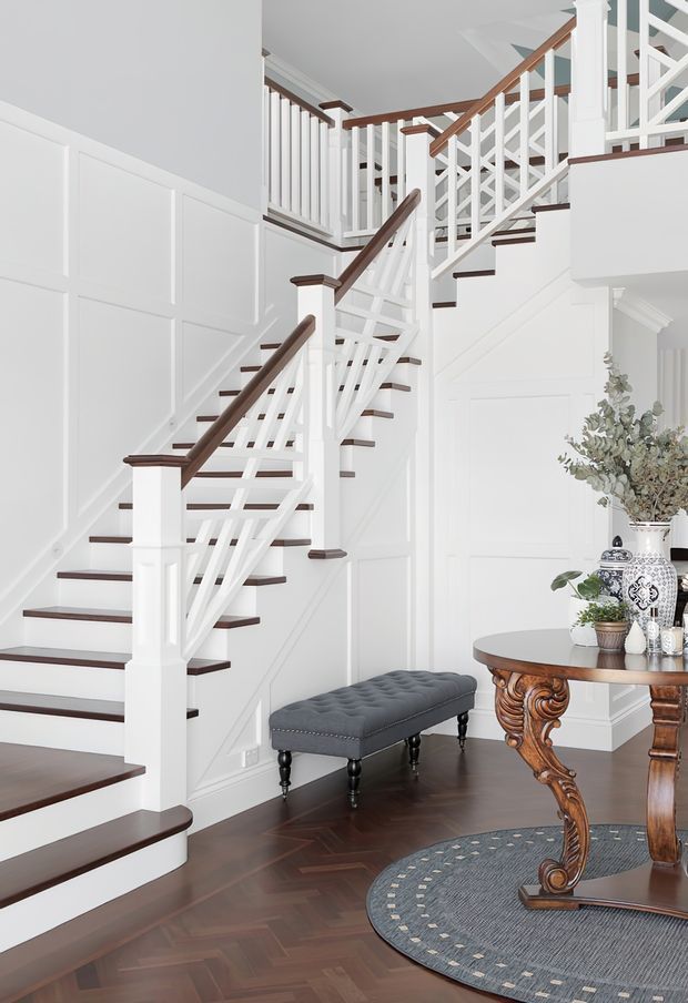 Bright, refined interior view of a multi-level stairway and landing. White painted wainscoted and panelled walls are paired with dark timber stair treads and handrails, white balustrades with decorative crossed detailing, and dark herringbone timber flooring. A tufted bench and ornate timber console table with decorative pottery occupy the entry area.