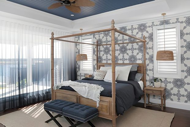 Bright, refined bedroom featuring a traditional four-poster timber bed with layered neutral bedding, a tufted bench, patterned blue-and-white wallpaper, plantation shutters, sheer floor-to-ceiling curtains, and a blue painted timber ceiling with a ceiling fan. The room has abundant natural light and appears to overlook a waterfront or open landscape through the glazing.