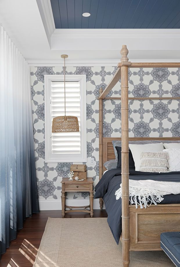 A bright, coastal-inspired bedroom featuring a substantial timber four-poster bed, patterned blue-and-white wallpaper, white plantation shutters, dark blue curtains, and a blue-painted tongue-and-groove ceiling. A woven pendant light, timber bedside table, layered neutral bedding, and a natural fibre rug add warmth and texture.
