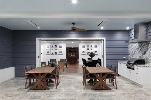 A spacious, covered outdoor entertaining area with two timber dining tables and chairs, opening into a bright interior living or dining space. The room features dark blue horizontal wall cladding, pale tiled flooring, a white paneled ceiling with recessed lights and ceiling fan, and a white outdoor kitchen with barbecue and dramatic marble-look splashback. Framed artwork and dark furnishings are visible in the adjoining room.