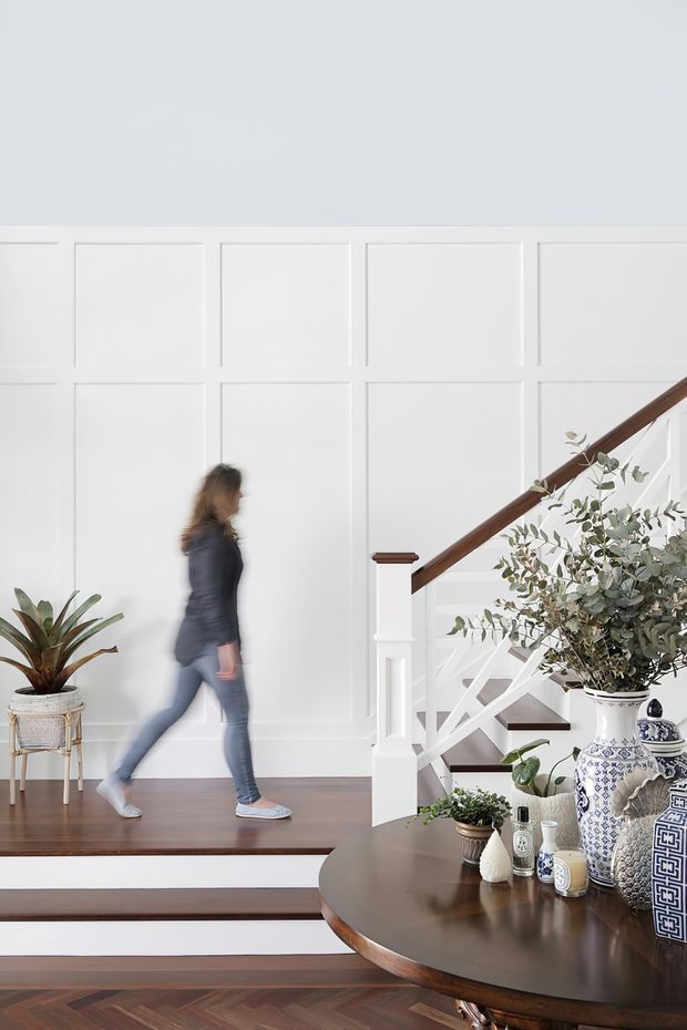 Bright, refined interior view of a staircase and split-level landing. White square-panel wall moulding forms the backdrop, with a dark timber handrail and white painted stair balustrade. Dark timber flooring and steps lead past a round dark wood table styled with blue-and-white ceramic vessels, a leafy branch arrangement, and potted plants. A blurred person is walking through the space.