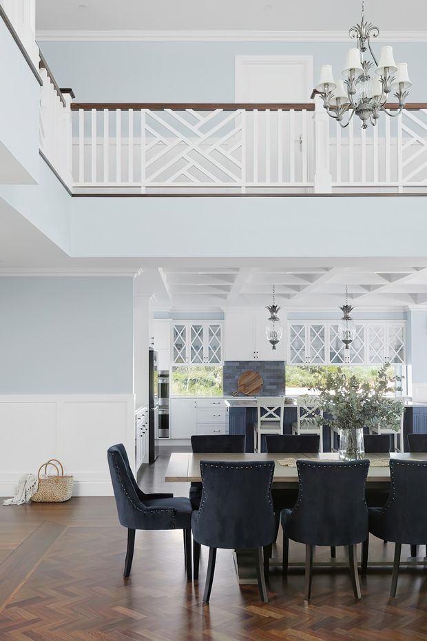 Bright, refined open-plan dining and kitchen interior with a dramatic upper-level balcony overlooking the room. The space features pale blue-grey walls, white wainscoting and trim, patterned white balcony balustrades, dark timber parquet flooring, a formal dark dining table with upholstered chairs, a white kitchen with cross-mullioned glass-front cabinetry, a decorative coffered ceiling, and ornate chandeliers.