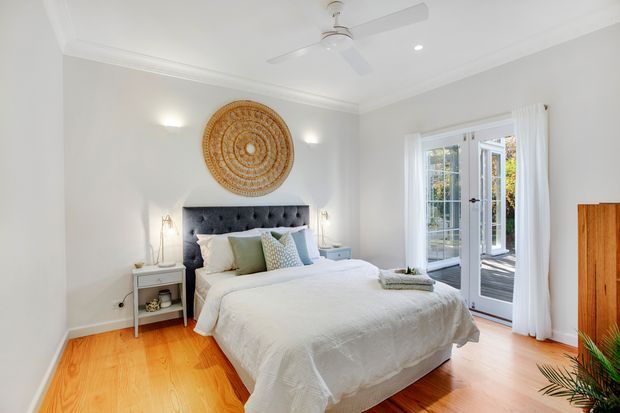 Bright, inviting bedroom with a large upholstered tufted headboard, white layered bedding, pale blue and neutral cushions, and matching bedside tables. The room has white walls with decorative cornices, light timber flooring, a ceiling fan, wall sconces, and full-height glazed French doors opening to an outdoor deck. A woven circular wall feature adds a warm natural accent.