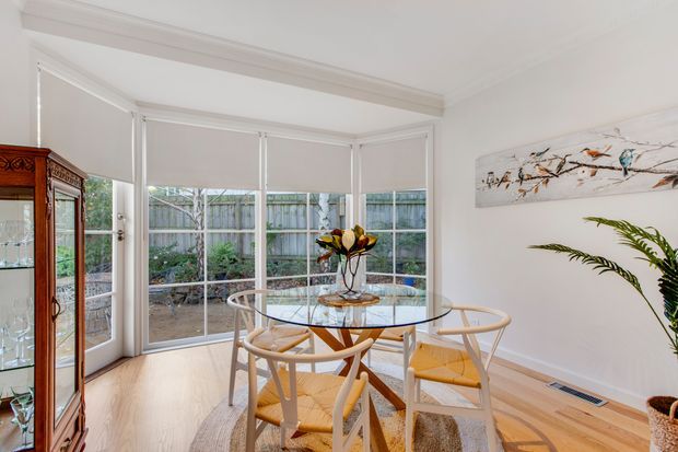 Bright dining area in a bay-windowed sunroom, featuring a round glass dining table with light timber and woven-seat chairs, pale timber flooring, white walls and trim, a traditional glass-front display cabinet, indoor plants, and artwork. The space opens visually to a fenced garden through expansive windows and glazed doors.
