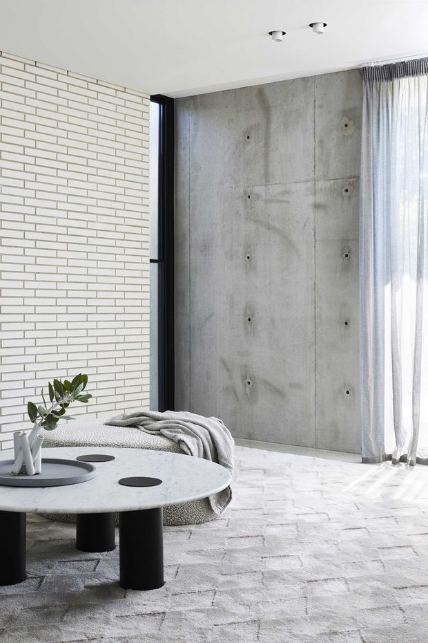 Bright contemporary living room with a restrained monochrome palette. The space features a pale brick feature wall, exposed concrete wall panels, black-framed glazing, sheer curtains, a light textured rug, and a low round marble or pale stone coffee table with black cylindrical legs. A neutral sofa with a draped throw and simple decorative objects completes the minimalist interior.