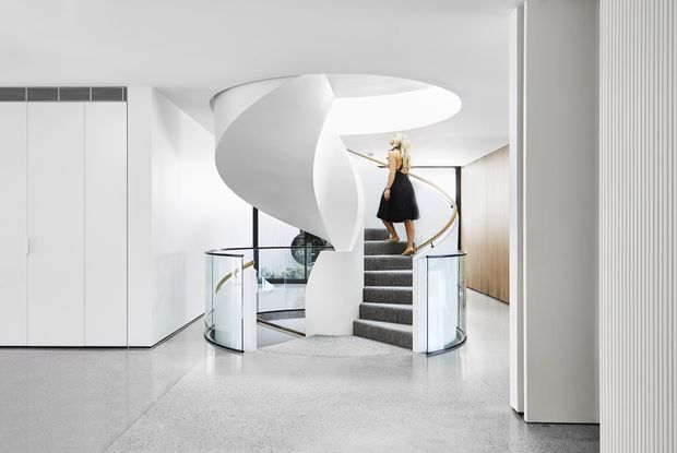A bright, highly contemporary interior centered on a sculptural white spiral staircase. The staircase has a smooth curved sculptural balustrade or central form, dark carpeted treads, clear glass guards, and a brass-toned handrail. The surrounding space features white walls and built-in cabinetry, a pale polished terrazzo-like floor, vertical wall panelling, and a double-height stair void. A person stands on the upper landing, but the architectural elements dominate the image.