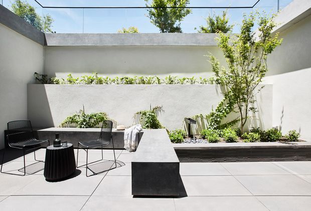 A bright, minimalist enclosed courtyard or terrace with pale rendered walls, large light paving slabs, raised planted beds, and a central built-in concrete bench or sculptural table. Black wire-frame outdoor chairs and a small black side table provide contrast, while a glass or open roofline brings in abundant daylight and greenery.