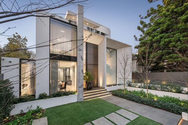 Contemporary multi-level residential facade viewed from a landscaped front garden, featuring crisp white rendered volumes, tall floor-to-ceiling glazing, a prominent concrete vertical element, warm timber-look entry cladding, broad entry steps, and large rectangular stepping pavers. The minimalist garden includes clipped hedges, a small lawn, and mature trees.