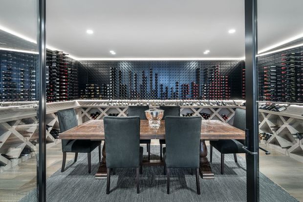 A sophisticated private wine room with floor-to-ceiling glass walls, extensive bottle storage lining the walls, and a central dark timber dining table surrounded by upholstered dining chairs. Integrated cross-braced wine racks and cool recessed ceiling lighting create a refined entertaining and tasting space.
