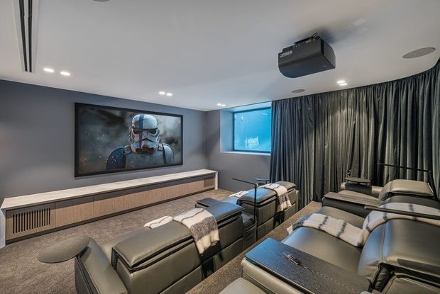 A sophisticated home cinema with multiple rows of reclining black leather lounge chairs, blankets, a large wall-mounted display showing a film, a ceiling-mounted projector, dark acoustic-style curtains, recessed lighting, and integrated media cabinetry. The room has a dark, immersive and comfortable atmosphere.