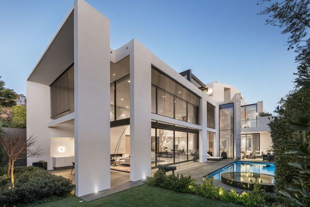 Contemporary multi-level residence with a sculptural white-rendered facade, tall projecting wall planes, extensive floor-to-ceiling glazing, and horizontal louvred screens. The house opens onto a landscaped courtyard with a narrow swimming pool and reflective water feature, visible through the transparent ground-floor living areas.