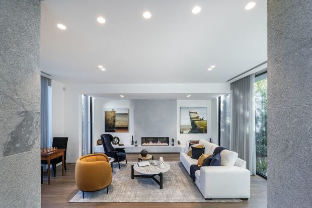 A bright contemporary living room viewed through a wide opening, with a white sectional sofa, mustard accent chair, dark lounge chairs, a low coffee table, rug, recessed ceiling lights, a linear fireplace, grey textured walls, and tall glazed openings with sheer curtains. A small dining or work area is visible to the left.