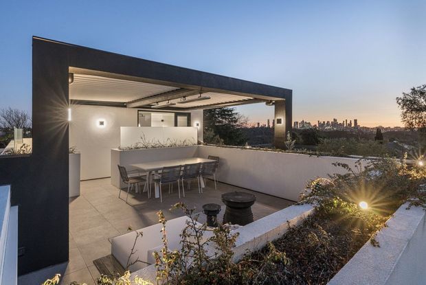 A contemporary rooftop terrace at dusk, framed by a dark rectangular architectural structure with a covered outdoor dining area. The terrace features a large dining table, built-in white rendered planters, tiled or paved flooring, integrated lighting, and landscaped garden beds, with a distant urban skyline visible beyond.