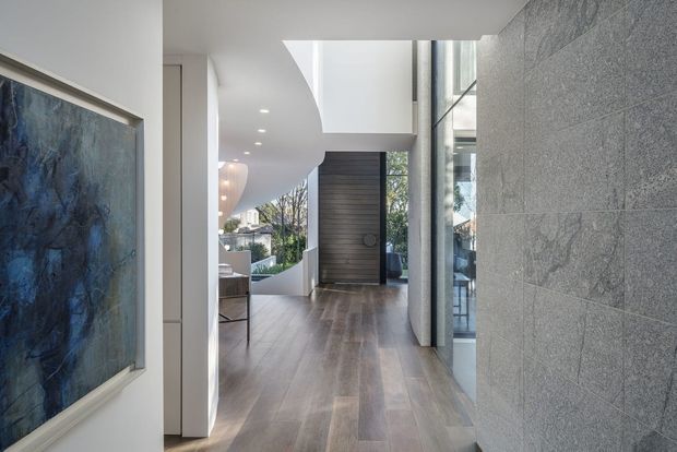 A bright contemporary residential entry hall with wide timber flooring, a tall dark timber pivot door, full-height glazing, recessed ceiling lights, and a dramatic curved white ceiling. A textured grey stone feature wall lines the right side, while a large abstract artwork is visible on the left.