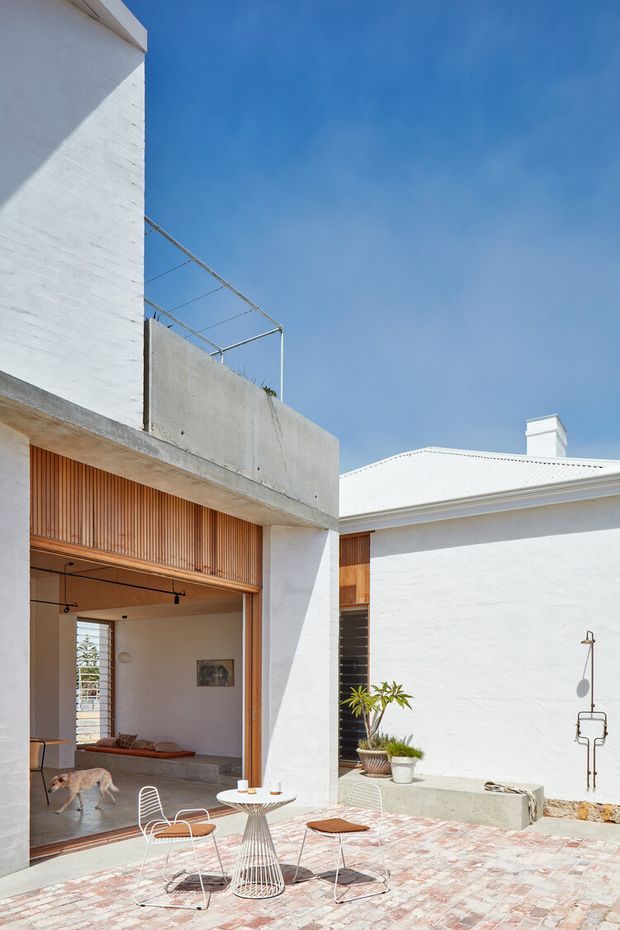 Sunlit courtyard of a white-rendered contemporary residence, with a paved outdoor seating area, small round table and wire-frame chairs. The courtyard is framed by a modern concrete volume with vertical timber screening and a more traditional white-roofed building with a chimney. An open glazed doorway reveals part of the interior, while potted plants and a dog add incidental detail.