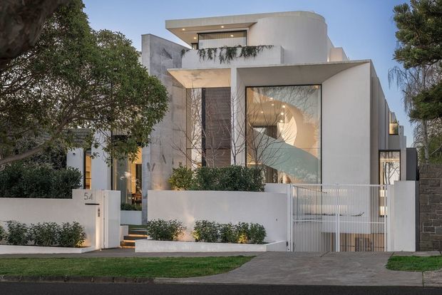 Street-facing view of a refined multi-storey contemporary residence with crisp white rendered walls, projecting box forms, curved upper-level geometry, large glazed openings, and a prominent illuminated spiral staircase visible through the front window. The property includes a low planted boundary wall, white metal driveway gate, landscaped frontage, and a mixed masonry side boundary.