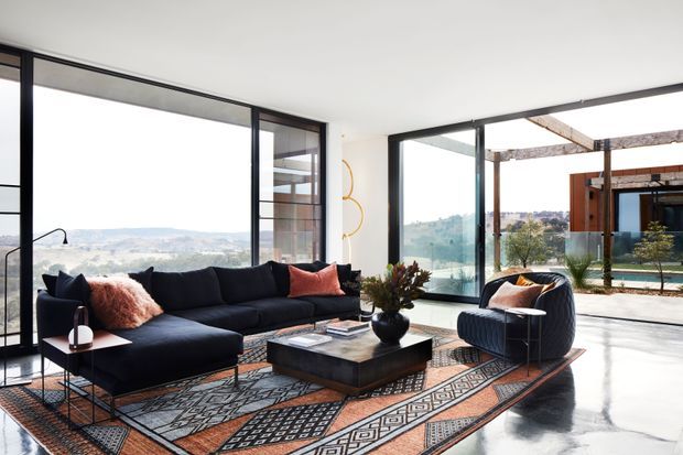 A bright, contemporary living room with a large dark sectional sofa, patterned area rug, sculptural coffee table, and an accent lounge chair. Full-height black-framed glazing opens views toward a landscaped outdoor area and distant rolling hills, while exposed timber pergola elements are visible outside. The space has polished dark flooring, white walls, and a refined indoor-outdoor connection.