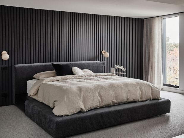 A contemporary bedroom with a low-profile upholstered platform bed, charcoal grey headboard and base, and layered neutral bedding. The room features a dramatic black vertical slatted feature wall, soft grey carpet, sheer curtains, and a large corner window bringing in natural light. Minimal bedside lighting and restrained styling create a refined, moody atmosphere.