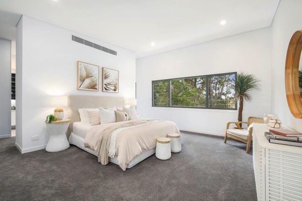 Bright, spacious contemporary bedroom with a large upholstered bed, soft neutral bedding, grey carpet, white walls, recessed ceiling lights, bedside tables, framed botanical artwork, a timber-framed lounge chair, and a wide horizontal window overlooking greenery. A louvred white cabinet and round timber-framed mirror are visible in the foreground.