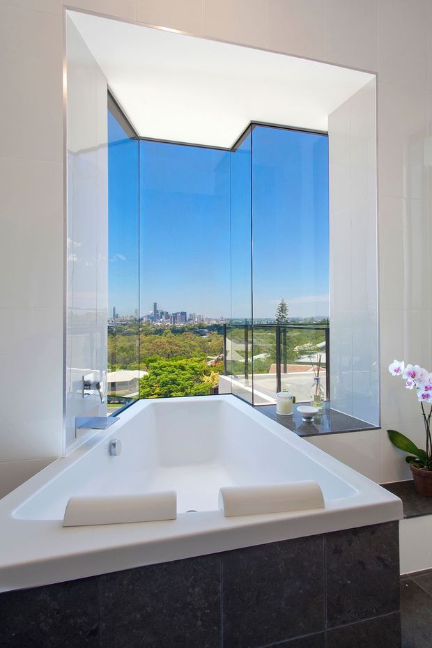 A bright, contemporary bathroom featuring a large freestanding built-in white bathtub positioned beside floor-to-ceiling corner glazing. The room has glossy white wall tiles, dark charcoal stone-look tiled bath cladding and ledges, and a panoramic urban and leafy outlook beyond the glass. A potted orchid and small bathroom accessories add restrained decorative detail.
